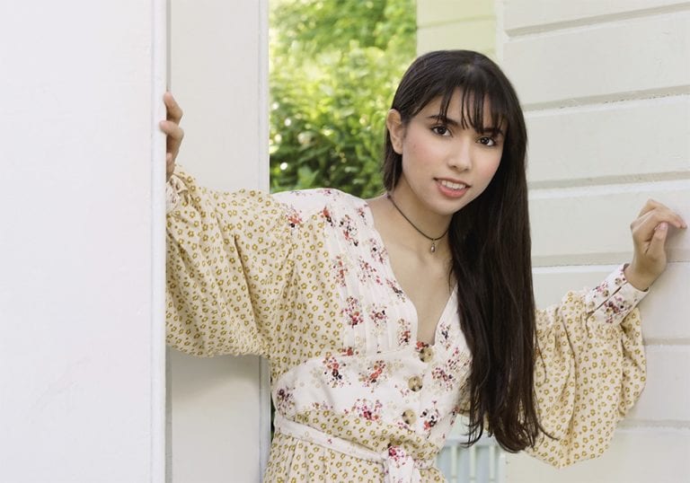 Portrait of a teen girl in a light dress leaning against a post with hands braced on two walls
