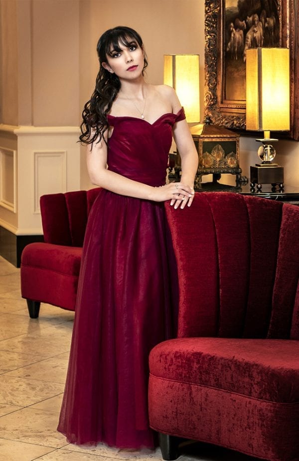 Portrait of a teen girl with a serious expression in an elegant dark red dress leaning against an red velvet chair
