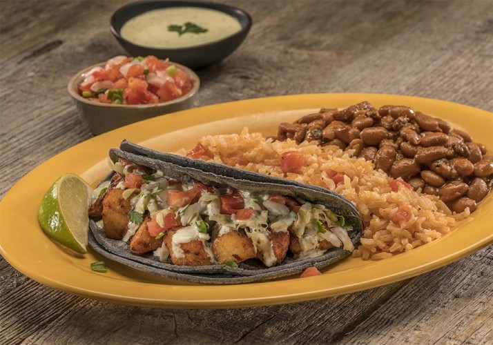 Black shelled taco on a plate with rice, beans, and a lime for seasoning