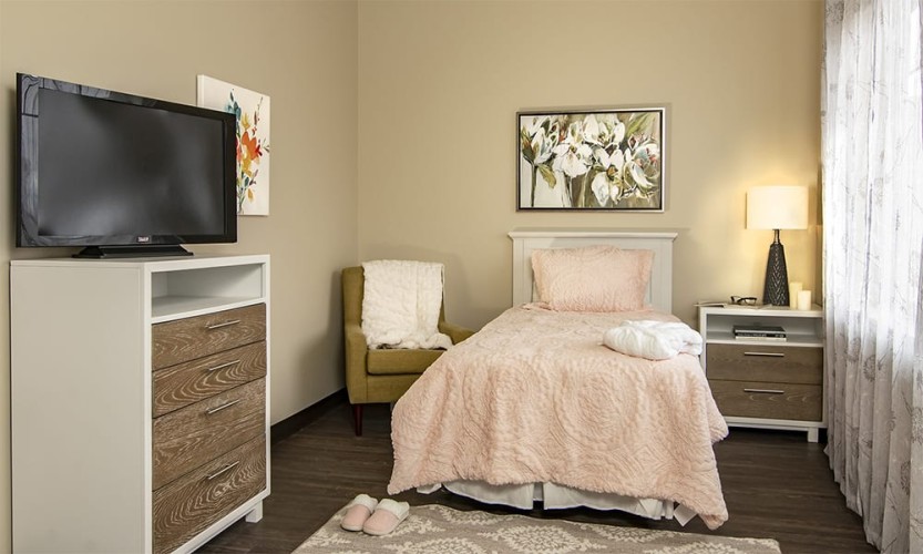 Small bedroom/living space with a bed covered in a light pink, fluffy comforter. Simple furnishings